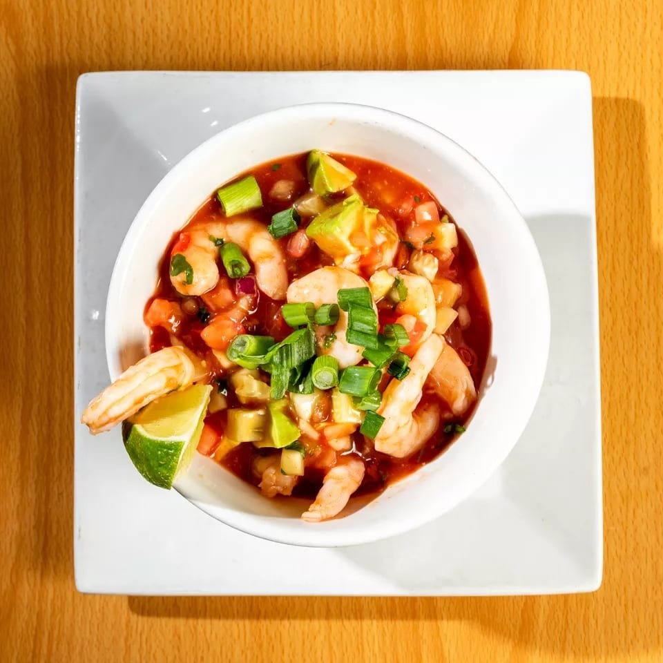Shrimp ceviche with avocado, tomatoes, and green onions in a white bowl on a wooden table.