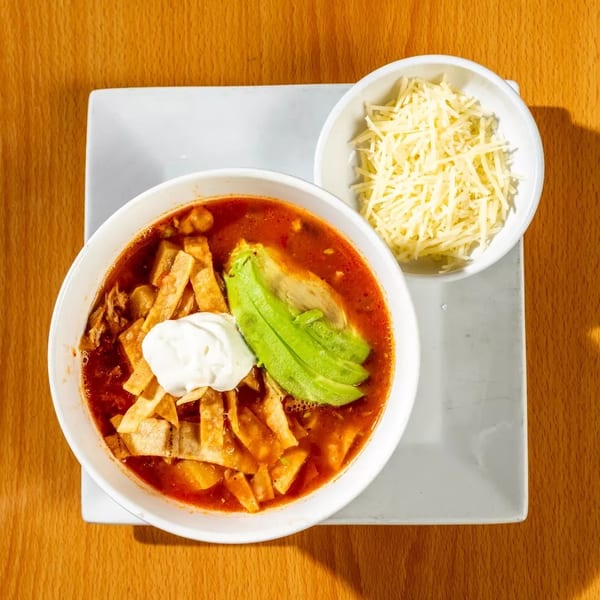 A bowl of tortilla soup with avocado, sour cream, and a side of shredded cheese.